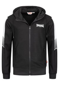 Fekete cipzáras kapucnis pulóver fehér csíkkal és csuklópánton cikkcakk mintával, Lonsdale London logóval a mellkason, valamint Union Jack címkével az oldalvarráson.