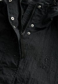 Giacca nera resistente all'acqua con zip frontale, bottoni a pressione e gocce sul tessuto testurizzato. Elementi distintivi includono una linguetta per il tiro.