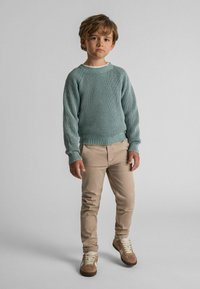 Lichtgroene gebreide trui met ribbeltextuur, beige aansluitende broek en bruine sneakers met witte veters. Staande pose tegen een neutrale achtergrond.