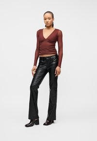 Γυναίκα με πλεξούδες, φορώντας εφαρμοστό μακρυμάνικο μπορντό τοπ με V λαιμόκοψη, μαύρο δερμάτινο παντελόνι με καμπάνα και σκούρα μποτάκια με τακούνι, όρθια.