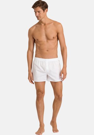 Man standing barefoot wearing white elastic-waist boxer shorts, looking down to his left on a plain light gray background.