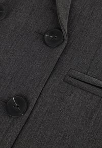 Harmaa bleiseri, jossa on tekstuuroitu kangas, kahdella mustalla napilla ja kapealla rintataskulla. Räätälöity muotoilu, jossa on siistit linjat.