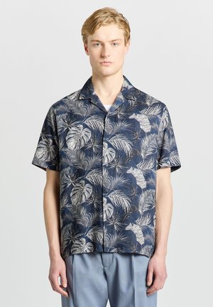 Homme blond portant une chemise bleu marine à manches courtes avec des motifs de feuilles tropicales blanches et grises et un pantalon bleu clair.