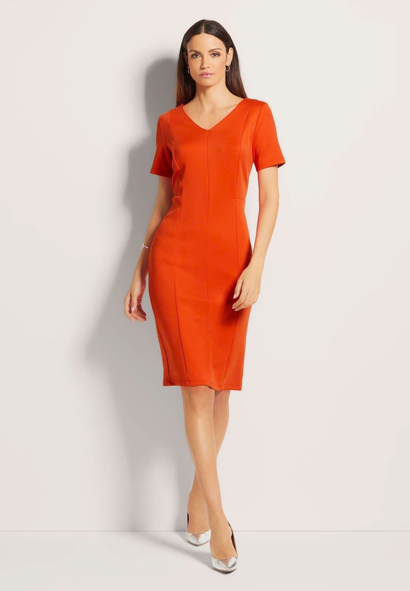 Rode aansluitende jurk met korte mouwen, V-hals, gestructureerde naden en een knielange zoom. Model gecombineerd met zilveren hakken, staand tegen een neutrale achtergrond.