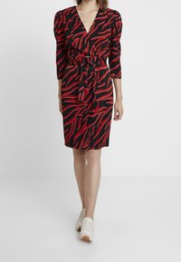 rotes und schwarzes Kleid mit V-Ausschnitt, Puffärmeln und tailliertem Bund. Es hat ein auffälliges gestreiftes Muster und einen knielangen Saum. Kombiniert mit weißen Sneakers.