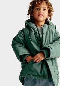 Zelená prošívaná bunda s kapucí, má patentky, boční kapsy a žebrované manžety, nošena přes šedý top a džíny.