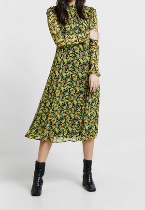 Kvinde iført en langærmet, blomsterprint midi-kjole med grønne, orange og gule mønstre, kombineret med sorte ankelstøvler, stående mod en ensfarvet baggrund.