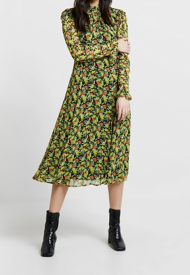 Femme portant une robe midi à manches longues avec des motifs floraux verts, orange et jaunes, assortie à des bottines noires, debout devant un fond uni.