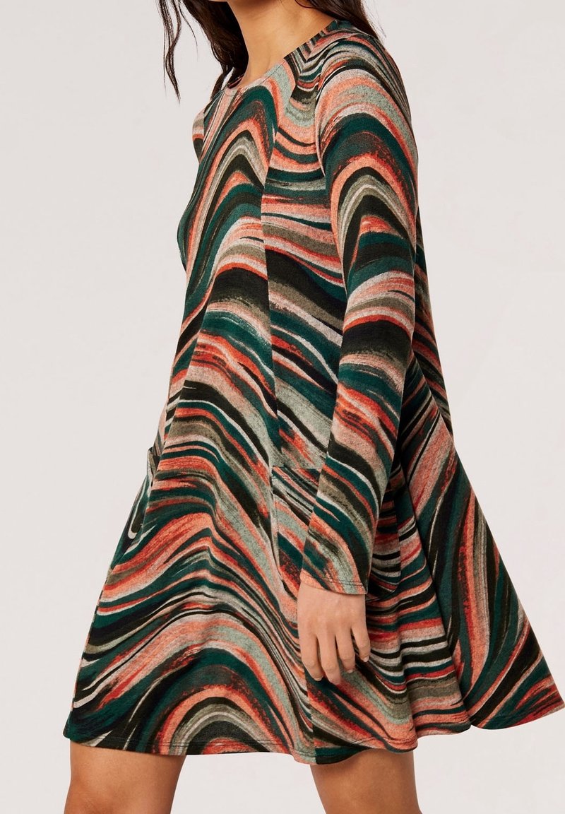 Femme portant une robe à manches longues en ligne A avec des motifs multicolores tourbillonnants dans des tons verts, orange, noir et beige.