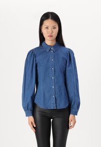Camisa de denim azul con mangas largas abullonadas, cuello puntiagudo y cierre frontal de botones. Presenta un dobladillo ligeramente curvado y una textura suave.