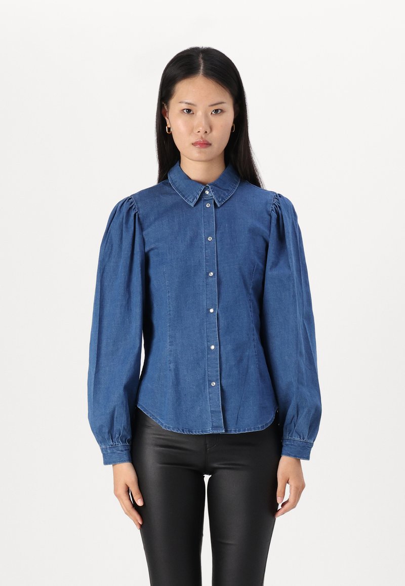 Camisa de denim azul con mangas largas abullonadas, cuello puntiagudo y cierre frontal de botones. Presenta un dobladillo ligeramente curvado y una textura suave.