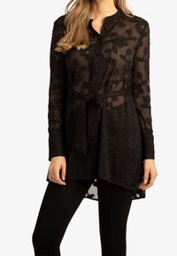 Donna che indossa una tunica nera trasparente a fiori con maniche lunghe e una cintura in vita sopra leggings neri, in piedi davanti a uno sfondo bianco.