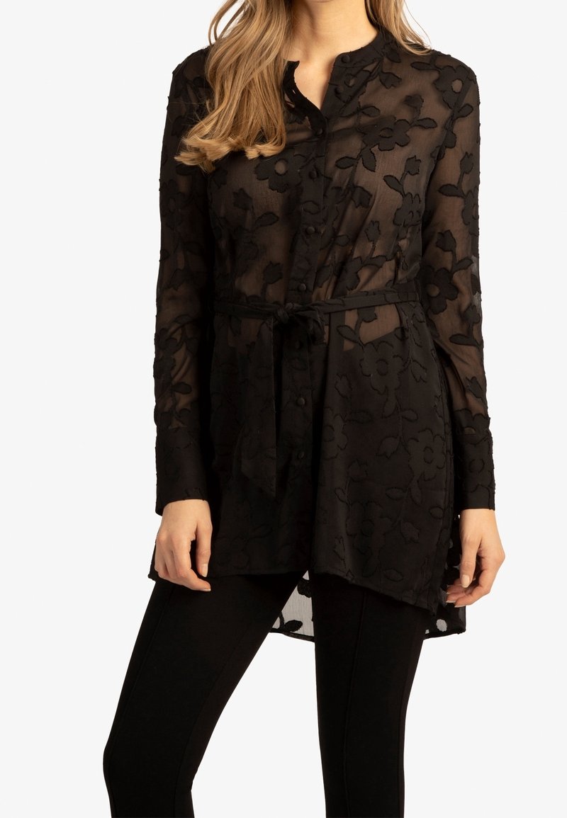 Donna che indossa una tunica nera trasparente a fiori con maniche lunghe e una cintura in vita sopra leggings neri, in piedi davanti a uno sfondo bianco.