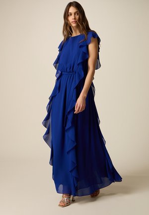 Donna che indossa un vestito lungo blu reale fluente con bordi arricciati e vita stretta, in piedi davanti a uno sfondo neutro.