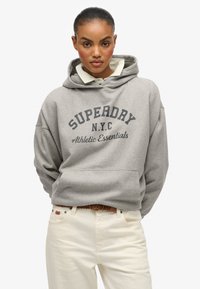 Joven con el cabello recogido que lleva una sudadera gris "Superdry N.Y.C Athletic Essentials" sobre una camisa blanca de cuello y pantalones crema.