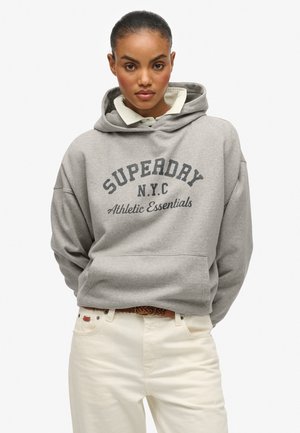 Jonge vrouw met opgestoken haar, die een grijze "Superdry N.Y.C Athletic Essentials" hoodie over een witte blouse en crèmekleurige broek draagt.