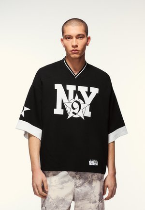 Young man with short hair wearing a black oversized shirt with white "NY" and star graphic, paired with light gray patterned pants.