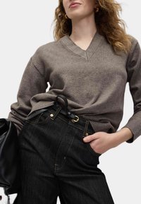Pull en laine marron à col en V associé à un jean denim noir taille haute, avec des accessoires dorés et une finition texturée. Sac noir inclus.