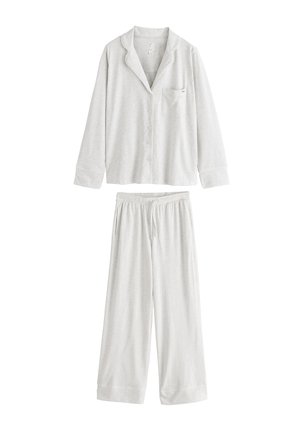 Vrouw met kort haar die een lichtbeige, knoopsluitend lang mouwen topje draagt met kraag en borstzak, staand voor doorschijnende gordijnen.