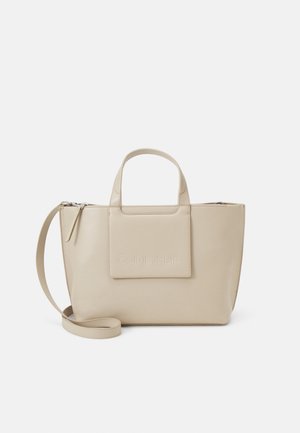 Beige synthetische handtas met dubbele bovenhandvatten en een afneembare schouderriem. Heeft een geperst logo op een voorste patch en een ritssluiting.