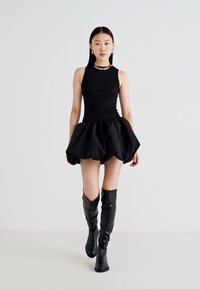 Vestido negro sin mangas con un corpiño ajustado y una falda voluminosa, combinado con botas de cuero negro hasta el muslo. Textura suave, diseño minimalista.