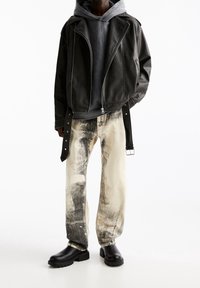 Personne portant une veste en cuir noire sur un sweat à capuche sombre, un jean décoloré blanc et noir, et des bottes noires épaisses sur un fond blanc.