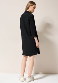 Abito camicia nero in tessuto strutturato, con maniche lunghe e colletto. Abbinato a sneakers crema, presenta una vestibilità rilassata e un orlo all'altezza del ginocchio.