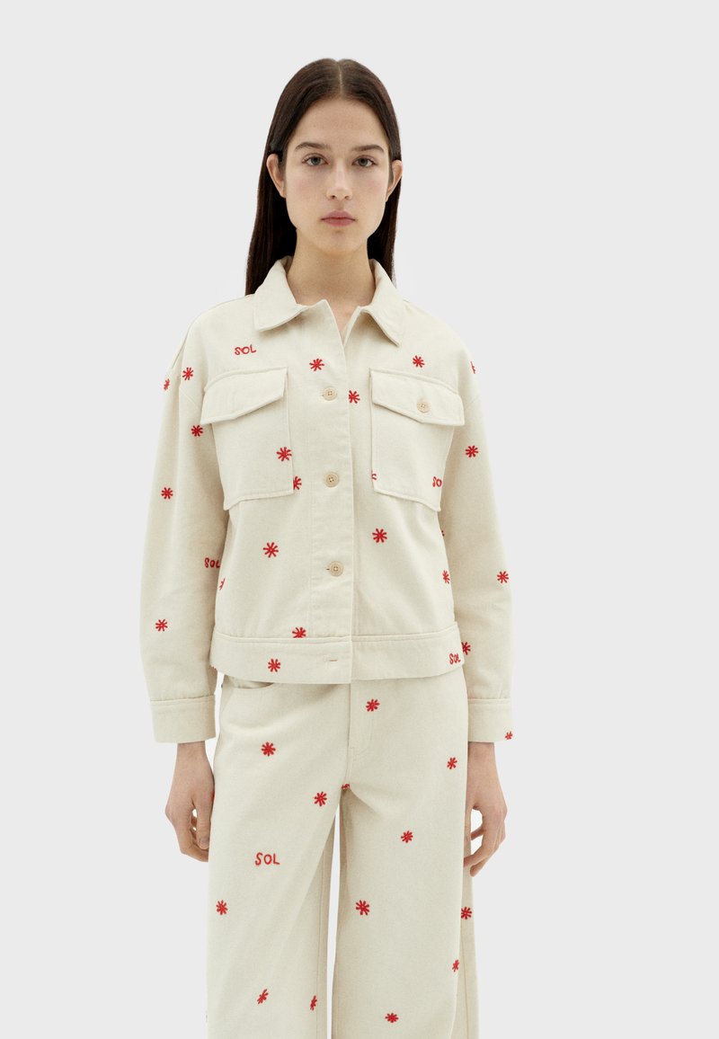 Roomwitte spijkerjas met rode bloemenborduursels, met twee borstzakken, knoopsluiting en een getailleerde taille. Gecombineerd met bijpassende broek.