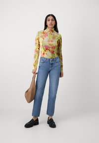 Langærmet gennemsigtig bluse med et farverigt blomstermønster i pink og grøn, parret med lyseblå lige jeans og sorte loafers.