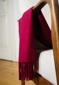 Écharpe en laine rouge foncé avec franges drapée sur le dossier d'une chaise beige tapissée à cadre en bois.