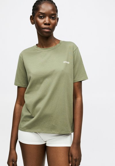 Femme portant un t-shirt HUGO vert olive et un short blanc, debout devant un fond blanc uni.