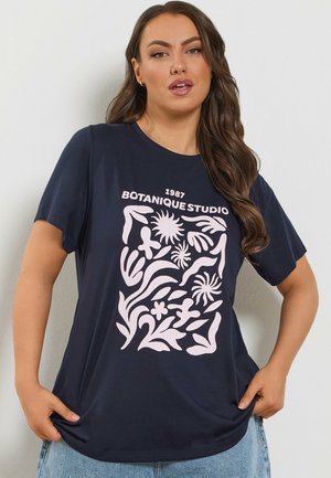 Kobieta z długimi falującymi włosami, ubrana w granatowy T-shirt z jasnoróżowymi abstrakcyjnymi kształtami roślinnymi i napisem "1987 Botanique Studio."