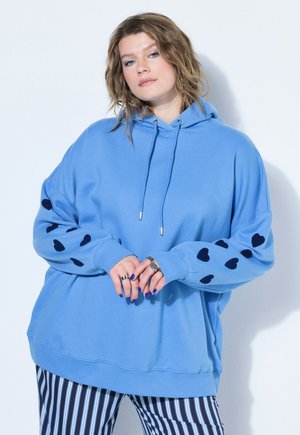 Frau mit welligem Haar, die einen hellblauen Hoodie mit schwarzen Herzen auf den Ärmeln und schwarz-weiß vertikal gestreifte Hosen trägt, steht vor einer weißen Wand.