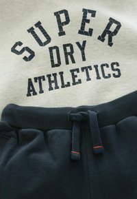 Grå sweatshirt med "SUPER DRY ATHLETICS" i revnet marineblå tryk over mørkeblå sweatpants med ribbet talje og snøre.