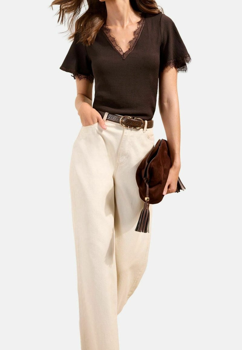 Femme portant un top marron foncé à col en V orné de dentelle, un pantalon large crème avec une ceinture tressée, tenant une pochette marron en daim avec pompons.