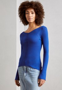 Blå ribbet langermet topp med en bred, asymmetrisk halslinje og sømdetaljer foran, sammen med et lys blått denimskjørt.