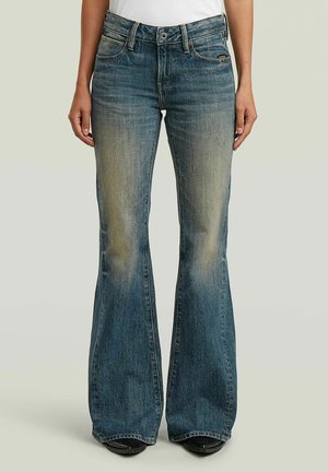 Personne portant un jean flare bleu délavé taille haute et des chaussures noires, debout devant un fond clair uni.