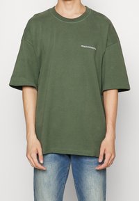 Camiseta de algodón de color verde oliva, de corte oversize, con mangas cortas y cuello redondo, que presenta un pequeño logotipo bordado en blanco en el pecho.
