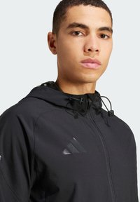 Schwarze Zip-Hoodie mit strukturiertem Finish, ausgestattet mit verstellbaren Kordeln, reflektierendem Logo und einem glatten, leichten Stoff.