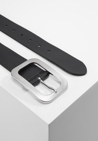 Ceinture en cuir noir avec une boucle rectangulaire en métal argenté, présentant une texture lisse et plusieurs trous d'ajustement le long de la sangle.