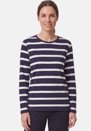 Frau lächelt, trägt ein marineblau-weiß horizontal gestreiftes Langarmshirt und dunkle Hose, steht vor einem schlichten weißen Hintergrund.