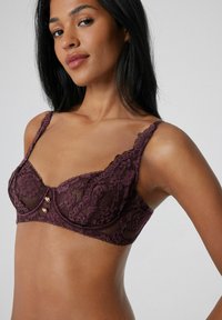 Krajková podprsenka v tmavě burgundské barvě s vlnitými okraji a květinovým vzorem. Obsahuje dekorativní knoflíky ve zlatém tónu uprostřed a nastavitelné ramínka.