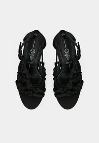 Pāris melnu augstpapēžu sandales ar siksnām, ziedu apdari un potīšu siksnām, attēlotas no augšas uz balta fona.