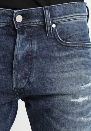 Zbliżenie na niebieskie wyblakłe dżinsy z metalowym guzikiem i niewielkimi przetarciami na udzie.