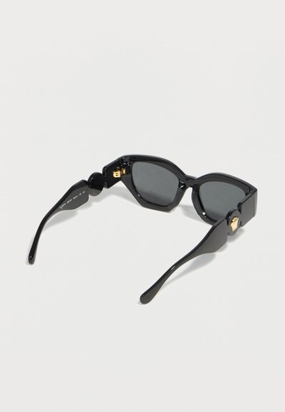 Μαύρα γωνιώδη γυαλιά ηλίου με σκούρους φακούς και χρυσές διακοσμήσεις με κεφάλι Μέδουσας στις πλευρές, που παρουσιάζονται ανοιχτά σε λευκή επιφάνεια.