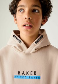 Sudadera beige con un collar alto, acentos en negro y un logo central que dice "BAKER BY TED BAKER" con un detalle en azul.