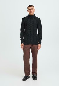 Maglione lavorato a maglia grigio scuro con colletto alto e motivo a trecce testurizzate, abbinato a pantaloni marroni e scarpe nere.