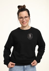 Leende kvinna med glasögon och hår i en hög knut bär en svart sweatshirt med en liten emblem-logotyp på bröstet och ljusblå jeans.