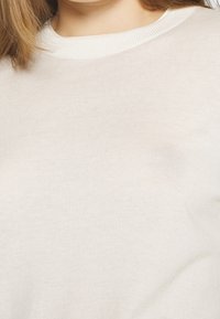 Světlý béžový pletený svetr s kulatým ribbovým výstřihem a hladkým povrchem. Detailní záběr ukazuje jemné steh a měkký, lehký vzhled.