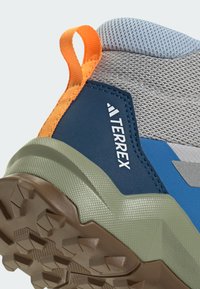Šedá a modrá sportovní obuv s oranžovým akcentem a texturovanou zelenou podrážkou; má síťovanou horní část a zesílenou patu pro podporu.